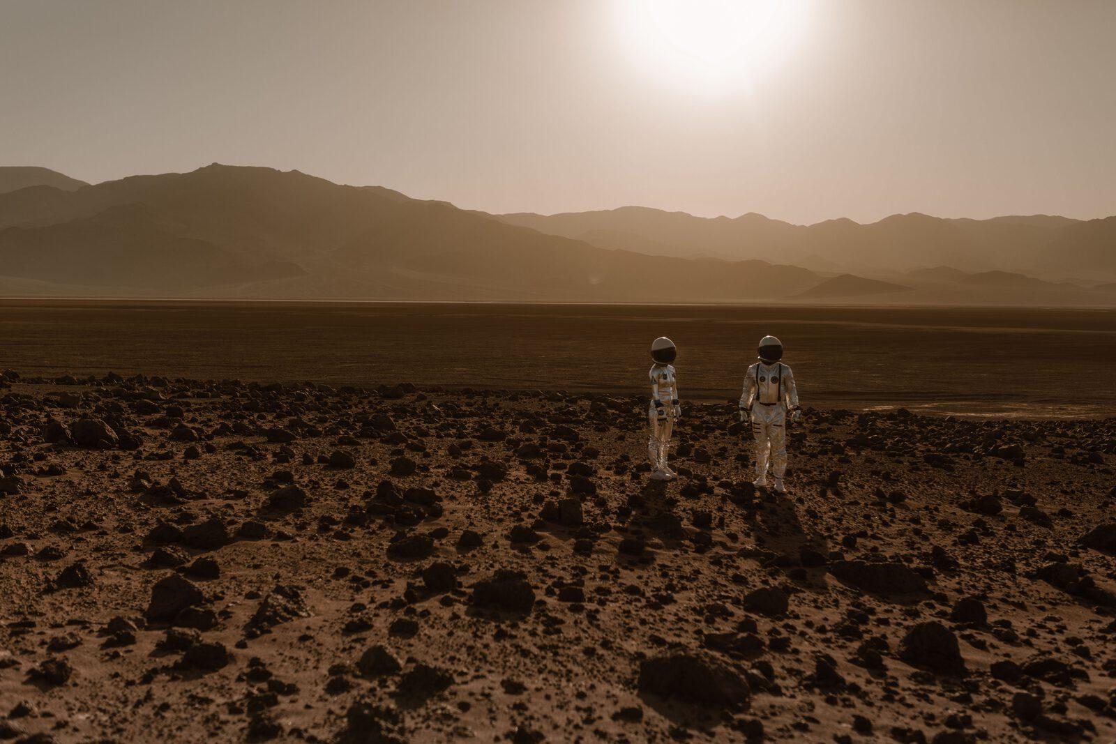 Two astronauts standing on a rocky Mars-like plain at sunset, mountains in the distance